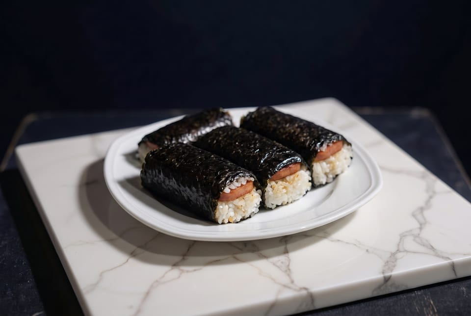 Four spam musubi, rice and spam wrapped in nori, on a white plate and marble board.