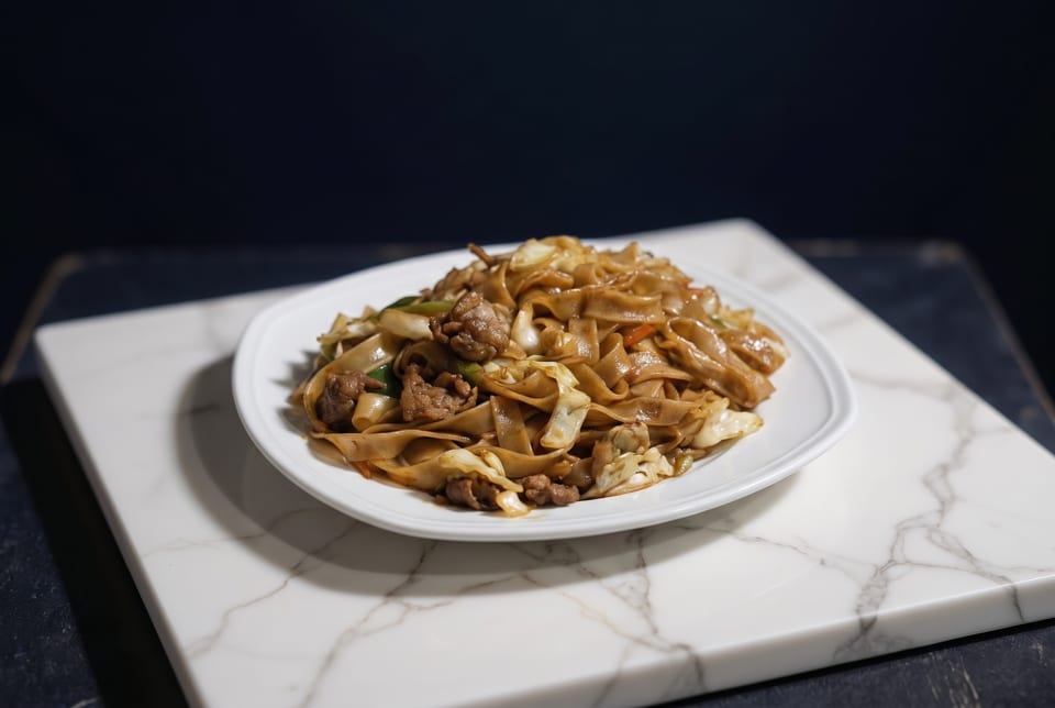 A white plate of stir-fried noodles with beef and vegetables on a marble slab.