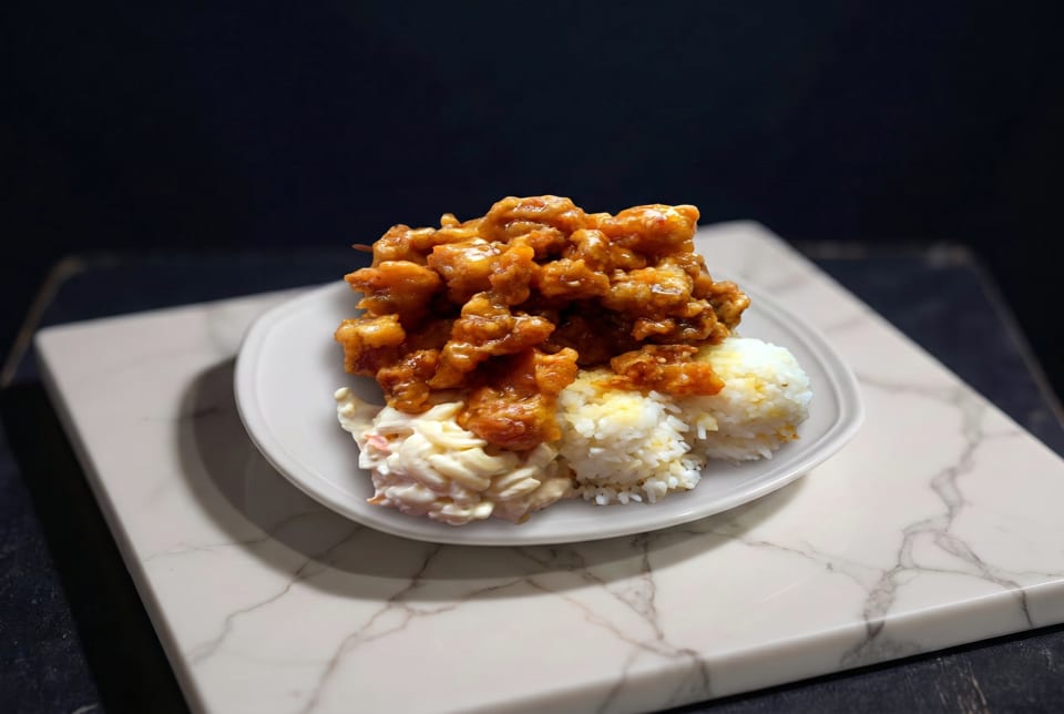 Orange chicken with white rice and macaroni salad on a white plate, on a marble slab.